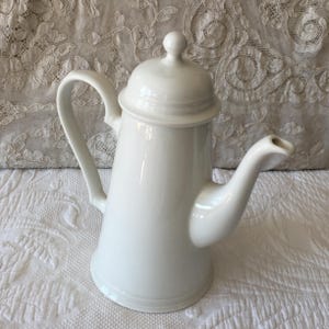 Könnte beinhalten: Weiße Kaffeekanne aus Keramik mit Deckel und Ausguss. Die Kanne hat einen runden Korpus und einen hohen, schlanken Ausguss.