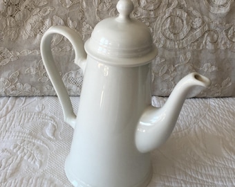 May include: White ceramic coffee pot with a lid and a spout. The pot has a rounded body and a tall, slender spout.