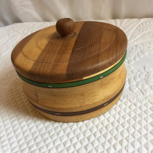 May include: A wooden storage container with a lid. The container is made of light and dark wood and has a green and brown trim. The lid has a knob on top.