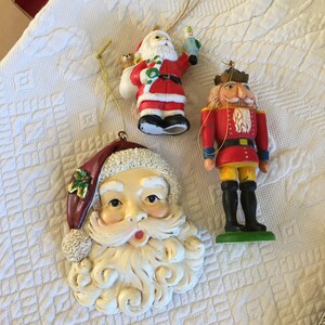 Könnte beinhalten: Drei vintage Weihnachtsbaumschmuck: ein Weihnachtsmann mit weißem Bart und rotem Anzug, ein Nussknacker mit rotem und gelbem Anzug und ein Weihnachtsmann-Gesicht mit weißem Bart.