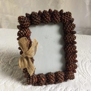 May include: A rectangular photo frame decorated with numerous small, brown pinecones. A tan burlap bow is attached to the lower left corner. The frame is designed to hold a photograph or artwork.