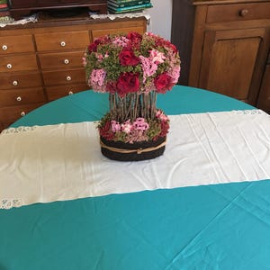 Puede incluir: Un arreglo floral con rosas rojas y flores rosas, sobre una base de ramitas y vegetación. Se encuentra sobre una mesa redonda con un mantel turquesa y un camino de mesa blanco con ribete de encaje.