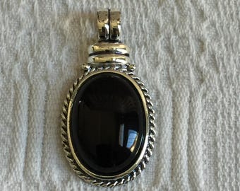 May include: Oval pendant with a polished black stone set in a silver-toned metal frame. The frame has a rope-like border and a loop at the top for a chain. The pendant is displayed on a white, textured surface.