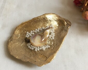 Puede incluir: Un plato de concha dorada con una concha iridiscente nacarada y una cadena de perlas blancas en su interior.