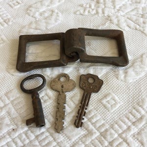 Könnte beinhalten: Eine antike Metallscharnier mit zwei rechteckigen Schlaufen und einem zentralen Zylinder sowie drei Vintage-Schlüsseln. Die Schlüssel sind in verschiedenen Formen und Größen, einer trägt die Nummer "6" und ein anderer den Namen "MASTER".