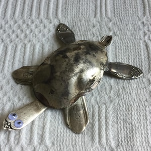May include: A metal turtle sculpture crafted from repurposed silverware. The turtle's body is a spoon, with forks and spoons forming the head, flippers, and tail. The flipper tips have small, light blue, painted eyes. The sculpture is set against a white background.