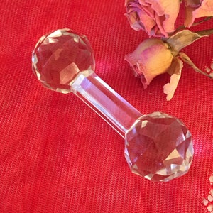 Puede incluir: Una mancuerna de cristal transparente con extremos facetados. La mancuerna está sobre una superficie de tela roja.