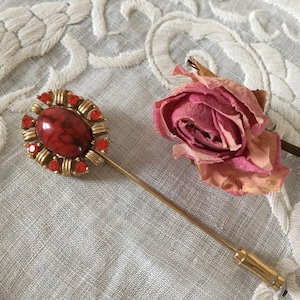 May include: A gold tone stick pin with a red stone surrounded by red rhinestones. The pin is on a white fabric background.