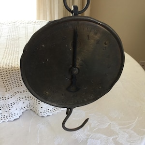 May include: Antique brass hanging scale with a black metal hook and a circular face with black markings. The face has numbers 1 through 8 and markings for 1/2 and 1/4 pounds.
