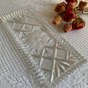 Könnte beinhalten: Ein klarer, rechteckiger Glasteller mit floralem und geometrischem Design. Der Teller hat ein rautenförmiges Muster in der Mitte und einen gezackten Rand. Getrocknete rote und hellbraune Rosen sind im Hintergrund.
