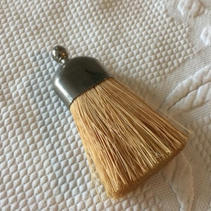 May include: A small, decorative brush with a silver-colored metal cap and a rounded top. The brush bristles are a light tan color. The brush is likely used for applying paint or other liquids.