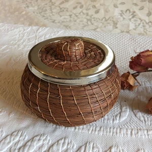 Pode incluir: Uma cesta redonda tecida com tampa. A cesta é feita de fibras naturais marrons e bege, com uma faixa de metal prateado ao redor da parte superior. A tampa tem um pequeno botão no centro. A cesta está em uma superfície branca texturizada.