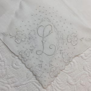 May include: A white handkerchief featuring a central embroidered letter L in a cursive script, surrounded by delicate floral motifs and small embroidered dots. The edges of the fabric are scalloped, and the entire design is monochromatic white.