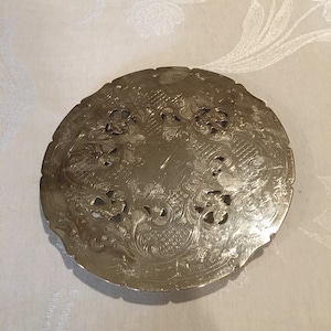 Puede incluir: Posavasos redondo plateado con un diseño intrincado y ornamentado. El posavasos tiene un borde elevado y un centro con un patrón floral.
