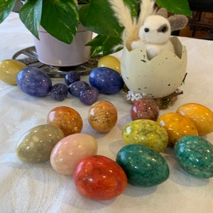 Puede incluir: Una colección de huevos de mármol decorativos en varios colores, incluyendo verde, amarillo, rosa, rojo y azul. Los huevos están dispuestos sobre un mantel blanco con un pequeño conejito de peluche sentado en una cáscara de huevo rota.