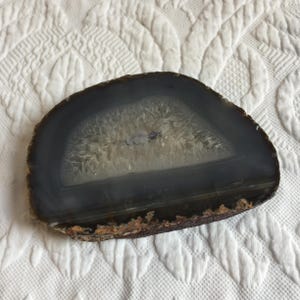 May include: A polished agate slice, showcasing a natural, semi-circular form. The outer edge is a dark gray, transitioning to a lighter, crystalline center. The bottom edge has a rough, brown texture. This decorative piece is ideal for home decor.