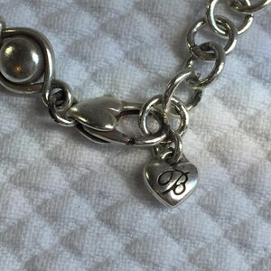 May include: A silver-toned bracelet featuring a series of circular links and a smooth spherical bead. The piece includes a heart-shaped lobster claw clasp and a small heart-shaped charm dangling from the clasp, which is engraved with the letter B.