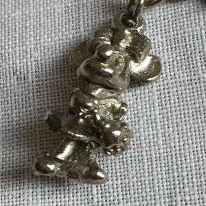 Może przedstawiać: Srebrny charms w kształcie postaci z kresk&oacute;wki, prawdopodobnie myszy. Charms jest szczeg&oacute;łowy z widocznymi rysami twarzy, uszami i ubraniem. Jest zawieszony na małej pętelce, odpowiedniej do bransoletki lub naszyjnika.