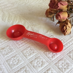 Op de afbeelding: Een rode plastic maatlepel met twee verschillende maten schepjes. De lepel heeft de woorden "Sunbrite It's Baiter" op de steel gedrukt.
