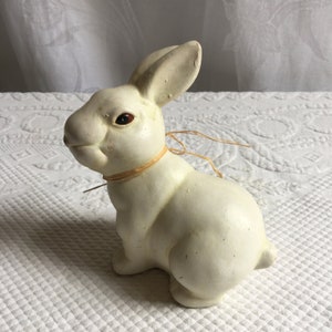 Puede incluir: Una figurita de conejo de cerámica blanca con ojos marrones y un lazo de paja alrededor del cuello. El conejo está sentado sobre un mantel blanco.