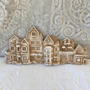 May include: A beige-colored, decorative wall hanging depicting a row of detailed buildings. The buildings include architectural details such as windows, doors, and awnings. The word "Boulangerie" is visible on one of the buildings.