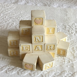 Puede incluir: Una pila de bloques de madera blancos con letras amarillas que deletrean "BEAR" y un corazón rosa en el bloque superior.