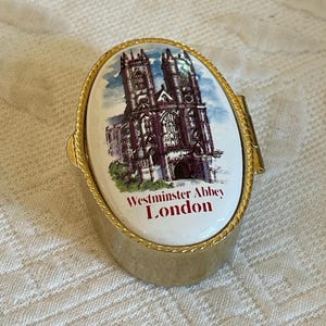May include: A gold-colored metal trinket box with a hinged lid. The lid features a colorful image of Westminster Abbey in London, with the text "Westminster Abbey London" printed below the image.