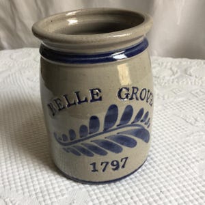 Pode incluir: Um pote de grés cilíndrico com acabamento cinza claro salpicado e borda azul escura. A frente apresenta o texto em relevo azul BELLE GROVE, um gráfico de folha de samambaia azul e o ano 1797 em relevo azul perto da base do recipiente.