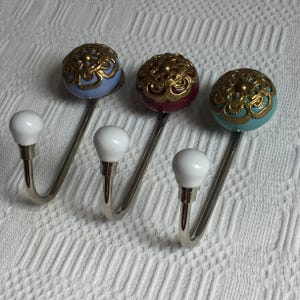 Peut inclure: Trois patères murales décoratives d'esthétique vintage. Chaque crochet comprend un bouton en céramique blanche, une tige incurvée en métal argenté et un orbe décoratif. Les orbes sont bleu clair, bordeaux et sarcelle, avec des détails en filigrane doré.