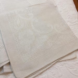 Könnte beinhalten: Nahaufnahme von gefalteten, cremefarbenen Leinen-Servietten. Jede Serviette hat eine detaillierte, strukturierte Bordüre und ein subtiles Blumenmuster auf der Oberfläche. Die Servietten sind gestapelt und zeigen das aufwendige Design und die Qualität des Stoffes.