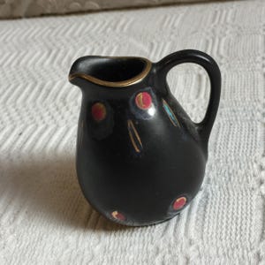 May include: A small, black ceramic creamer with a gold rim and handle. The creamer is decorated with red and gold circular accents and teal lines. The creamer is approximately 4 inches tall.