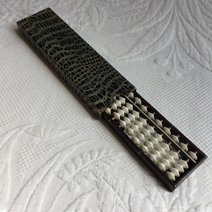 May include: A vintage abacus with white beads and a dark wooden frame, partially inside a black case with a reptile skin pattern. The abacus is a traditional calculating tool.
