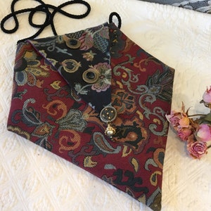 Könnte beinhalten: Eine kleine, dreieckige Handtasche mit einem schwarzen Riemen. Die Tasche ist aus einem roten, goldenen und blauen gemusterten Stoff gefertigt. Sie hat eine Klappe mit einem schwarzen Stoff darunter und vier dekorative Knöpfe.