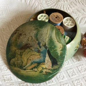 以下が含まれることがあります： 花柄の緑色の金属製のヴィンテージ裁縫箱。蓋には、庭園に座っている2人の人物が描かれた絵が描かれています。箱は開いており、糸巻きやその他の裁縫用品が入っています。