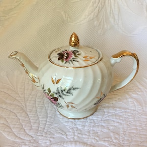 May include: White teapot with a gold trim and floral design. The teapot has a swirling pattern and a gold knob on the lid.
