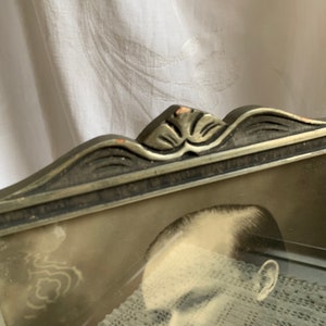 May include: A vintage silver-toned picture frame with an ornate design. The frame is slightly damaged on the top edge. The frame is holding a black and white photograph of a person's face.