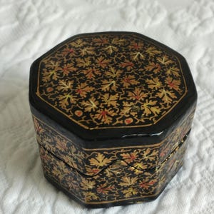 May include: An octagonal black box with a hinged lid. The box is decorated with a repeating pattern of gold, red, and yellow leaf shapes on a black background. The box is sitting on a white surface.