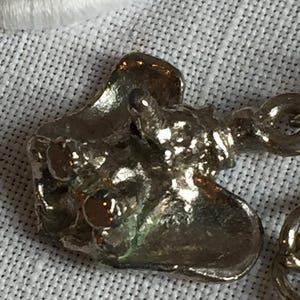 Może przedstawiać: Zbliżenie srebrnego charmsa o organicznym, teksturowanym wzorze. Charms ma zaokrąglony kształt z nieregularnymi krawędziami i małą pętelką do mocowania. Powierzchnia odbija światło, podkreślając jego szczeg&oacute;łowe wykonanie.