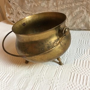 May include: A small, antique brass cauldron with a rounded body, three small feet, and a curved handle. The cauldron has a tarnished, aged appearance, with visible wear and tear. The interior is dark, and the handle is attached to the side with a small knob.