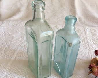 Puede incluir: Dos botellas de vidrio vintage con un tinte azul claro. Las botellas son de forma rectangular y tienen un aspecto ligeramente esmerilado.