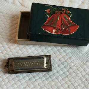 May include: A vintage Hohner harmonica with the brand name etched on the metal surface. The harmonica is next to an open box decorated with a Christmas bell design in red and green on a dark green background.