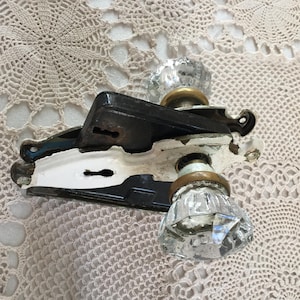 Op de afbeelding: Vintage deurbeslagset met een zwart en wit geschilderde metalen plaat met een sleutelgat en twee heldere, facetgeslepen glazen deurknoppen. De knoppen hebben messingkleurige metalen bases. De set vertoont gebruikssporen.