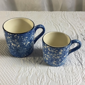 Puede incluir: Dos tazas de cerámica con un patrón moteado de esponja en azul y blanco. Cada taza tiene un interior de color crema, un borde azul oscuro y un asa curva. La taza más grande está situada a la izquierda, mientras que la más pequeña está a la derecha.