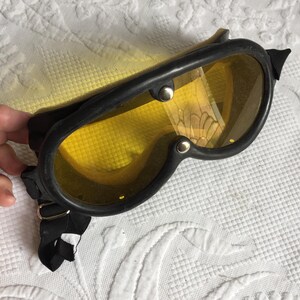 Può includere: Occhiali da moto neri vintage con lenti gialle. Gli occhiali hanno una montatura in gomma nera e un cinturino regolabile nero. Le lenti sono tenute in posizione da rivetti argentati. Gli occhiali sono appoggiati su una superficie bianca strutturata.