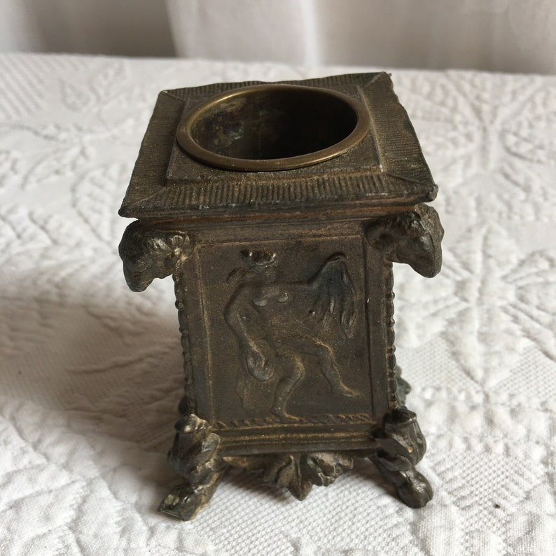 May include: A small, square, bronze inkwell with a decorative design of a winged figure. The inkwell has a round opening at the top and sits on four feet.