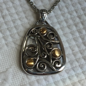 May include: A silver-toned pendant necklace featuring an openwork design with swirling metal patterns and three circular gold-toned accents. The pendant is suspended from a matching silver-toned chain link necklace with a decorative bail.