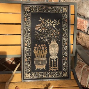 May include: Framed artwork featuring a black background with a detailed floral and vase design. The artwork includes a vase on a stand, a potted plant, and a branch with fruit. The frame is silver.