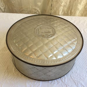May include: A round silver-colored metal tin featuring a quilted diamond pattern on the lid and sides. The center of the lid displays a rectangular embossed emblem with text. The container has a dark brown rim along the top edge.