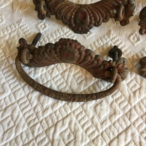 May include: A pair of antique, ornate, cast iron drawer pulls with a reddish brown patina. One pull is a simple, decorative, half-circle shape. The other pull is a more elaborate, oval-shaped pull with a curved handle.