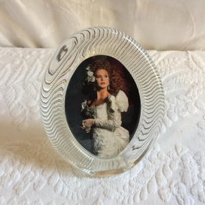 Könnte beinhalten: Ovaler Bilderrahmen aus Glas mit einem gewellten, strukturierten Design. Der Rahmen enthält ein Foto einer Frau in einem weißen Kleid mit einer großen Schleife.